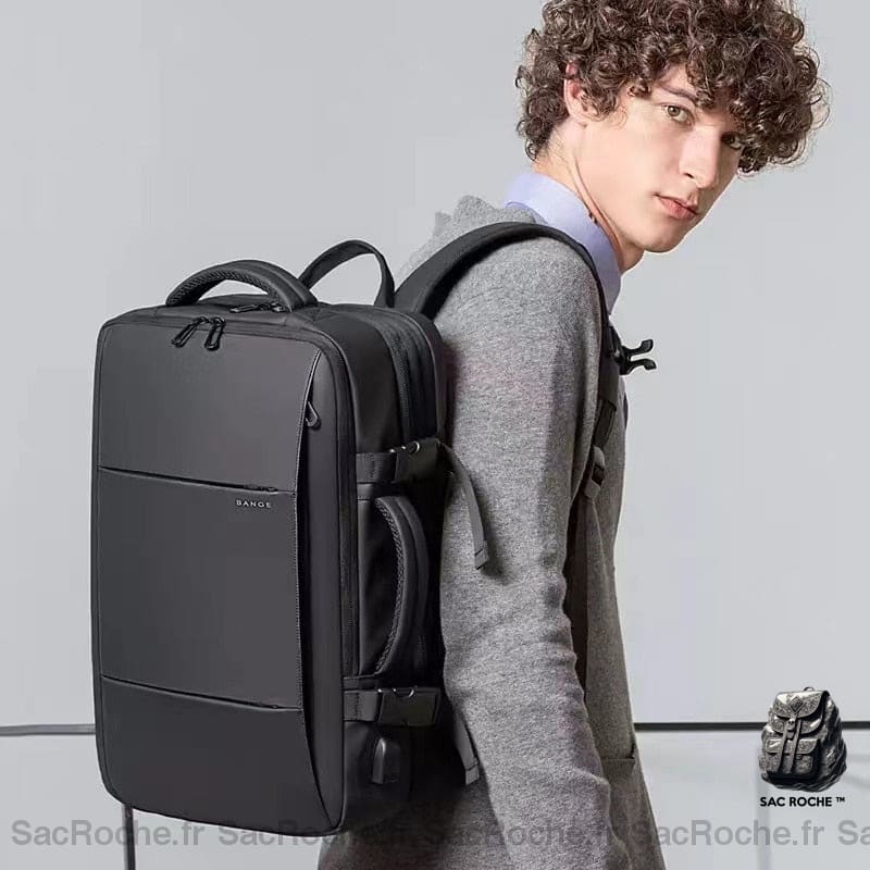 Un jeune homme portant à l'épaule un sac à dos valise rectangulaire noir. Celui-ci se ferme avec une glissière sur le dessus et a une hanse sur ses deux faces.